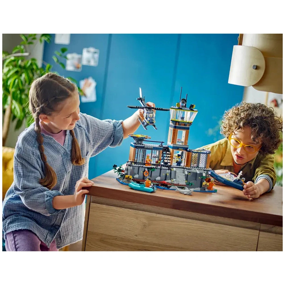 A girl with braided hair plays with a LEGO City Police Prison Island set, while a boy in glasses observes closely. The detailed prison features a control tower and various accessories, set amidst a colorful backdrop.