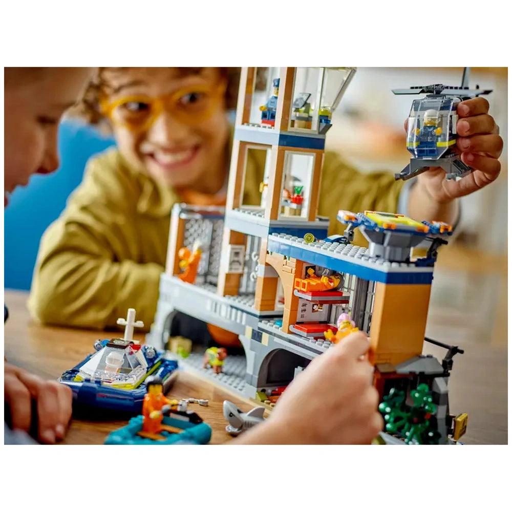 Two children engaged with the LEGO® City Police Prison Island set, smiling as they explore its detailed features. One child is holding a small helicopter, while the other focuses on the intricate cliffside prison. A toy boat and various colorful minifigures are scattered around the play area. Bright colors and imaginative designs encourage creative play and storytelling.