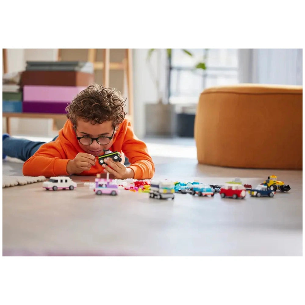 A child with curly hair wearing glasses explores a small black toy vehicle while lying on a light-colored floor, surrounded by colorful LEGO vehicle creations.