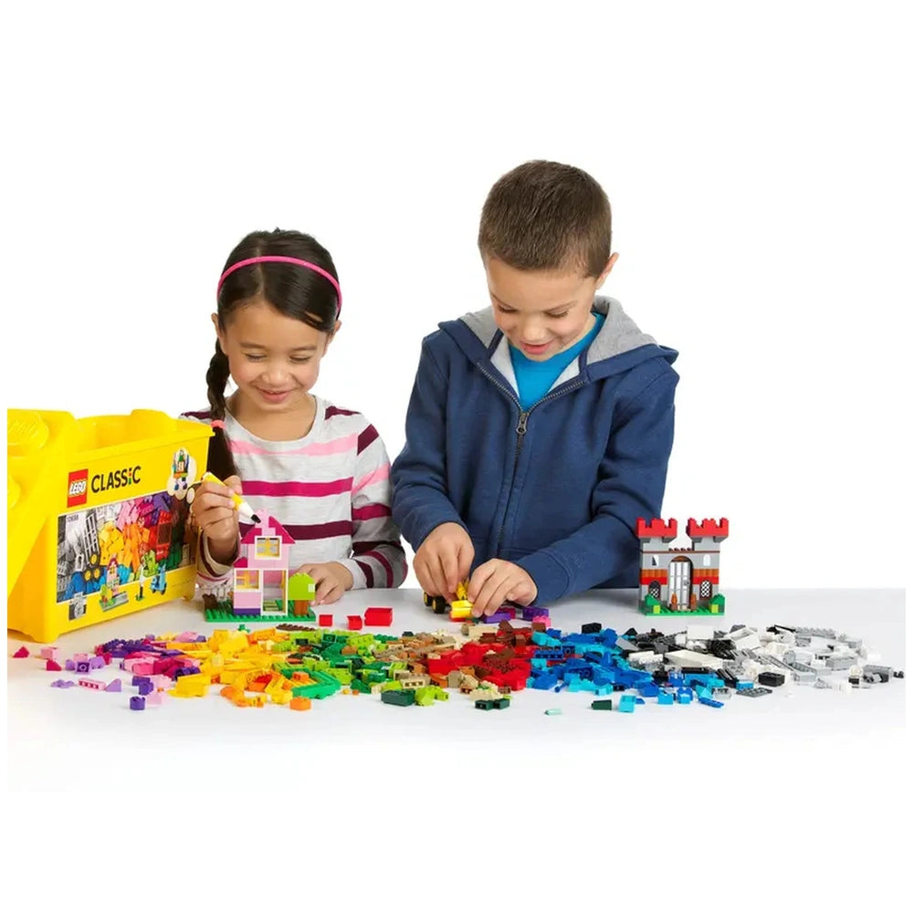 Two children engaged in building with colorful LEGO bricks spread across a table. A yellow LEGO Classic box is on the left, featuring various vibrant pieces. One child is assembling a pink house, while the other focuses on a small vehicle near a castle structure.