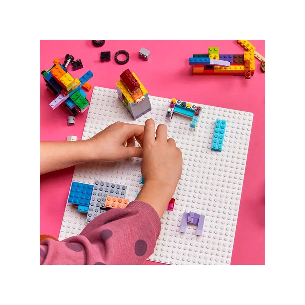 A child's hand builds with colorful LEGO bricks on a white baseplate, surrounded by scattered pieces on a pink surface.
