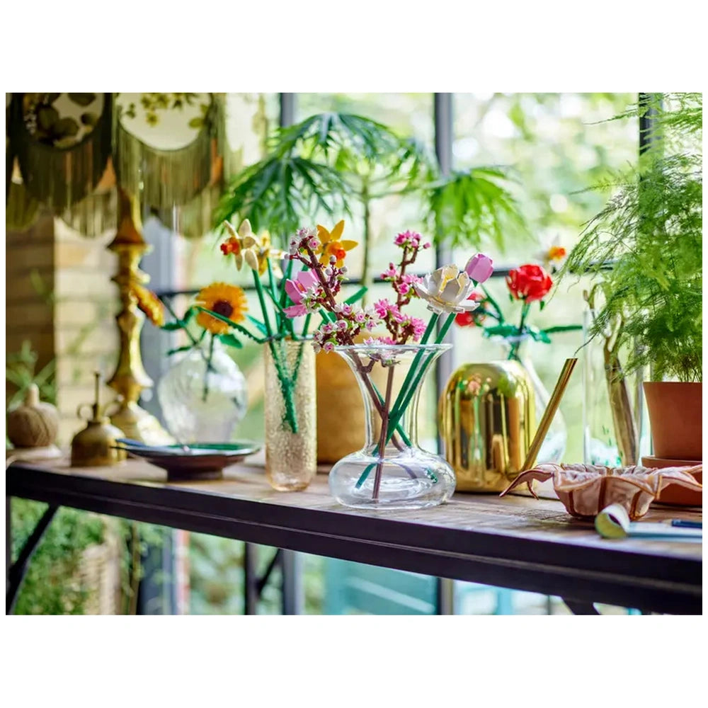 Colorful cherry blossom and flower arrangements in various vases on a wooden table, surrounded by greenery and decorative items.