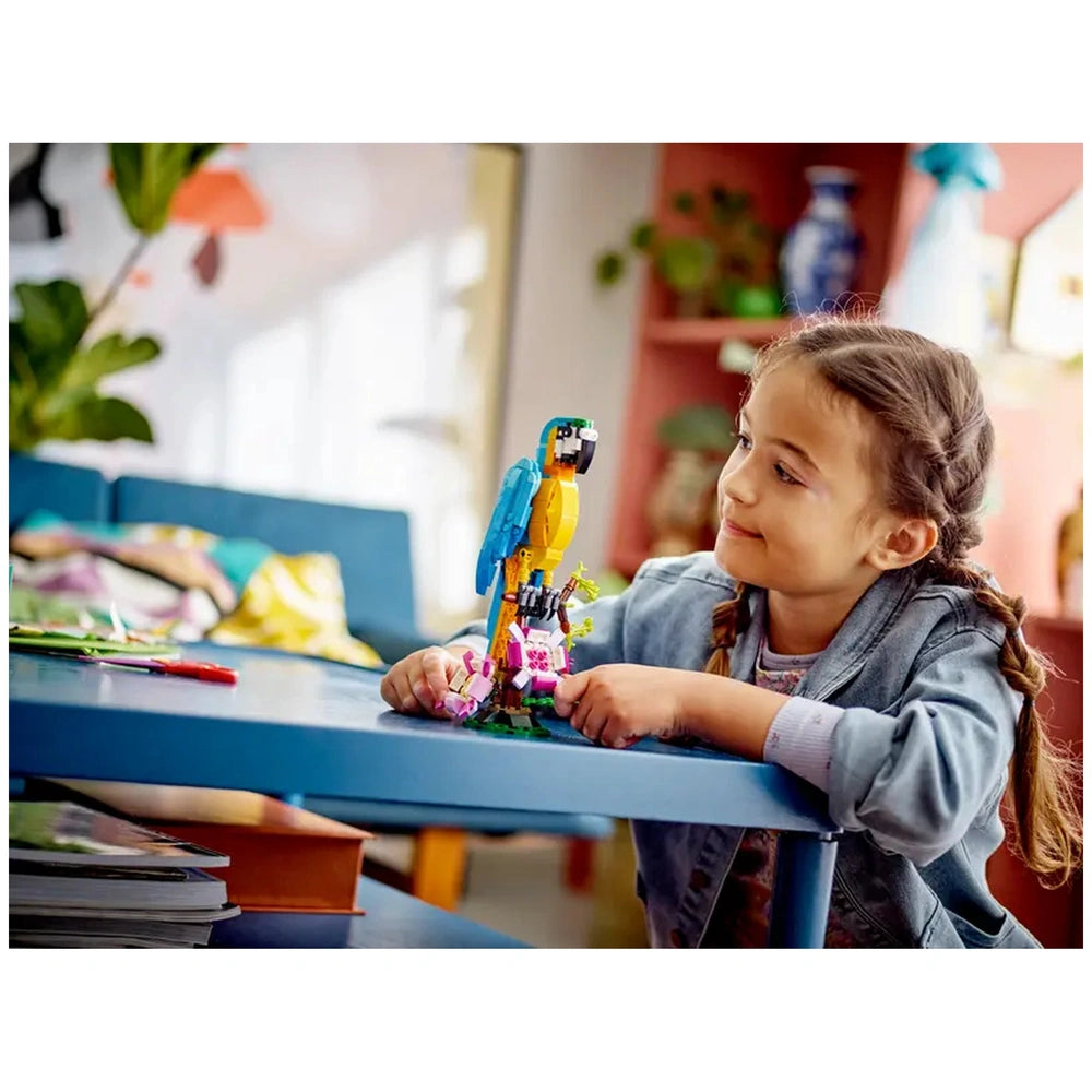 A young girl with braided hair smiles while playing with a colorful LEGO® parrot on a blue table, surrounded by vibrant decor.