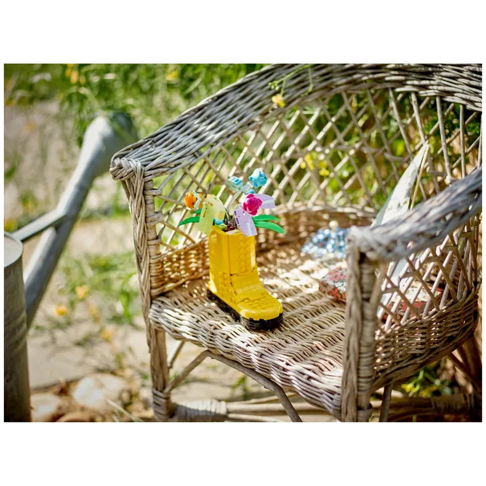 Yellow LEGO watering can shaped like a boot filled with colorful flower toys, placed on a woven chair surrounded by greenery.
