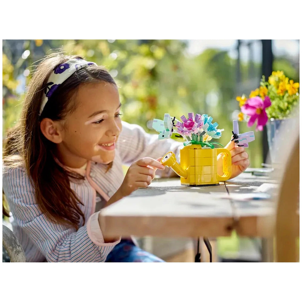 A young girl smiles while interacting with a vibrant Lego Creator 3in1 Flowers in Watering Can set on a wooden table. The cheerful yellow watering can has colorful flower toys with movable petals and delicate butterfly toys attached. A bright garden backdrop enhances the playful scene.
