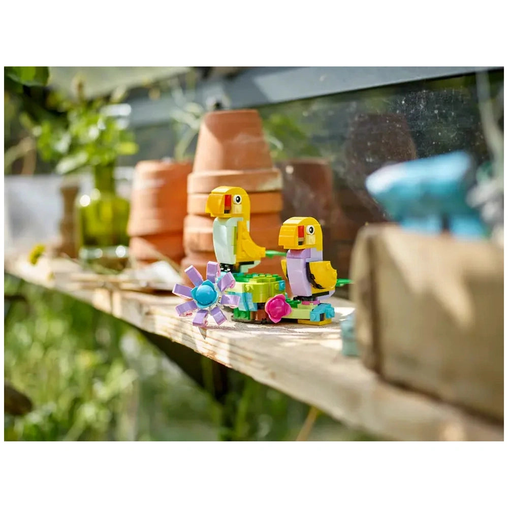Two colorful LEGO® bird toys with yellow heads and purple bodies, sitting beside a vibrant flower toy on a wooden shelf. Behind them, terracotta pots and greenery create a sunny garden ambiance.