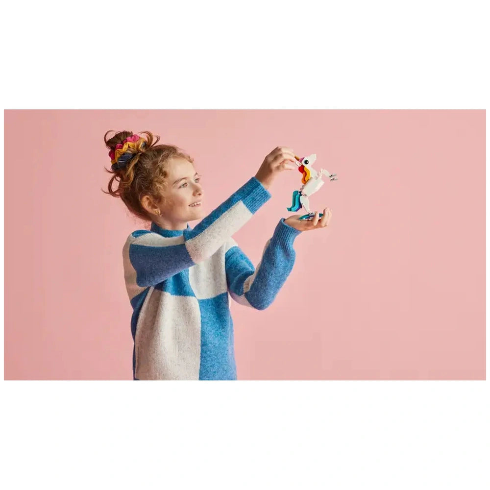 A smiling child with curly hair holds a colorful LEGO Creator Magical Unicorn, featuring a golden horn and vibrant tail, against a pink background.