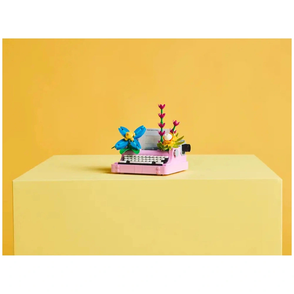 LEGO Creator Typewriter in pink adorned with blue and yellow flowers, set on a light yellow table against a vibrant yellow background.
