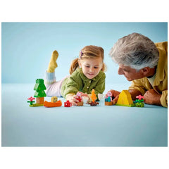 A young girl smiles while playing with a vibrant LEGO® DUPLO® Camping Trip set, featuring Peppa Pig figures, a canoe, and colorful accessories. An older man leans in with interest, sharing the playful experience. They are surrounded by a light blue background, creating a cheerful, engaging atmosphere.