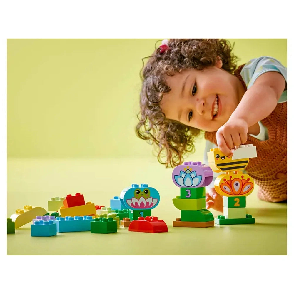 A joyful toddler with curly hair smiles while stacking colorful LEGO® DUPLO® bricks, including flowers and playful animal figures, on a bright green surface.