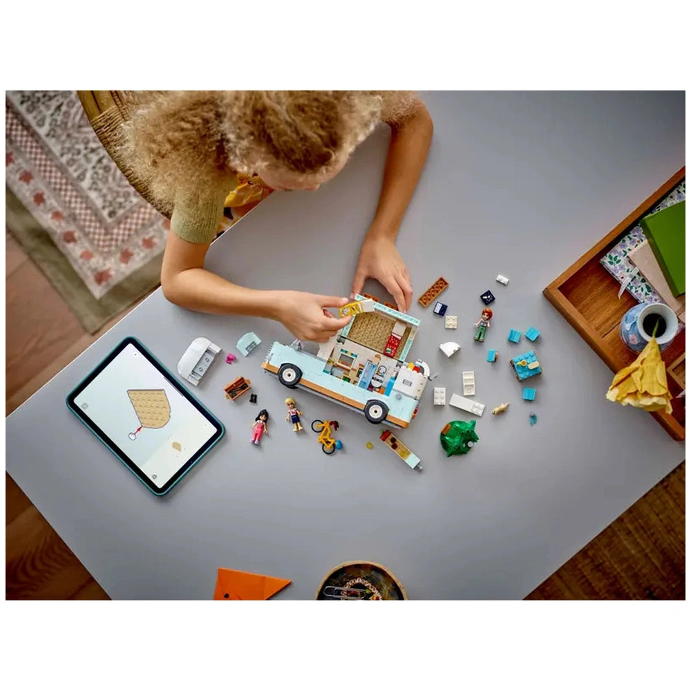 A child plays with the LEGO® Friends Friendship Camper Van, placing accessories inside the open van's colorful interior. Surrounding the camper are minidolls, a ferret figure, and colorful building pieces, with a tablet displaying building instructions nearby.