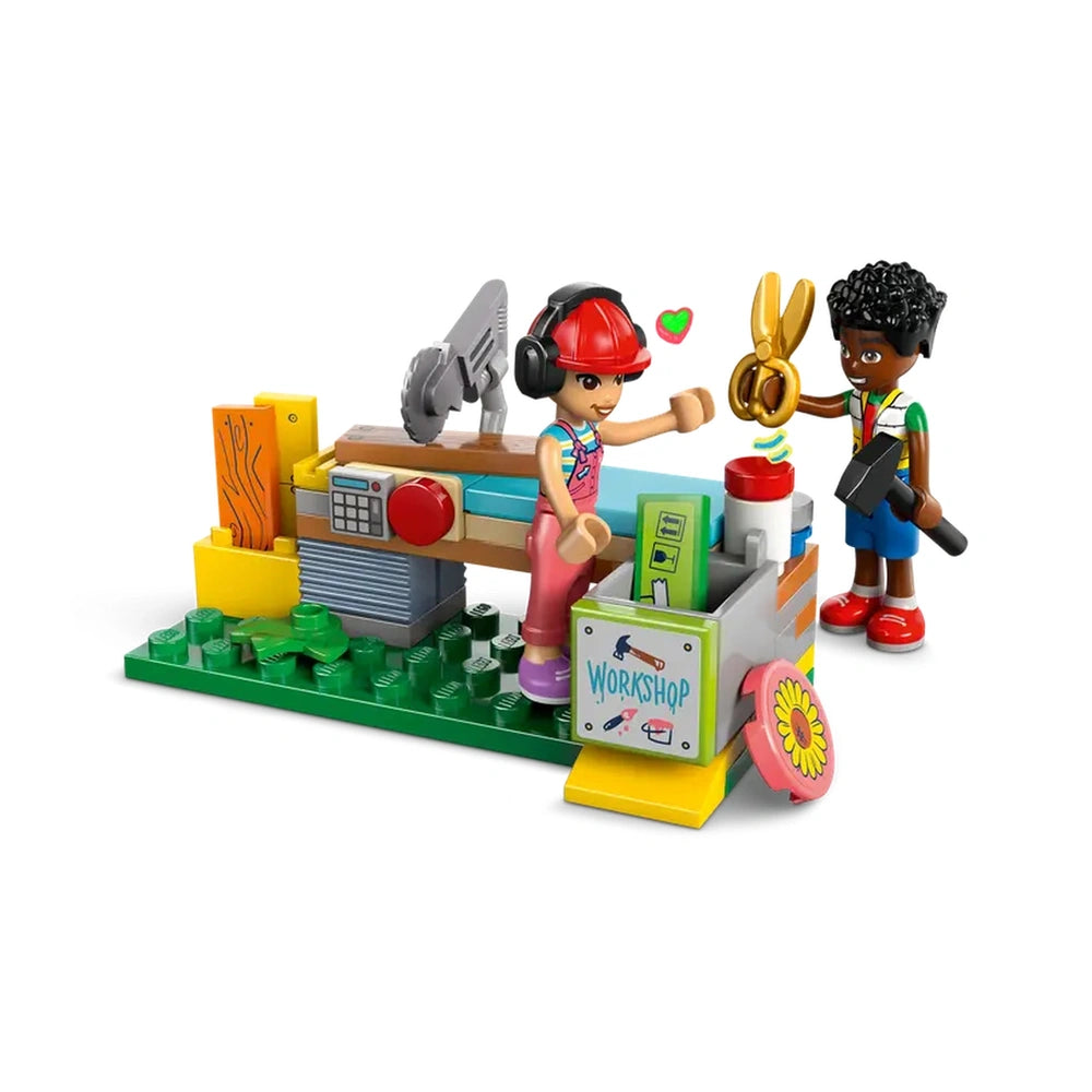 Two LEGO Friends minidolls at a colorful workshop, one holding scissors and the other with a hammer, surrounded by tools and greenery.