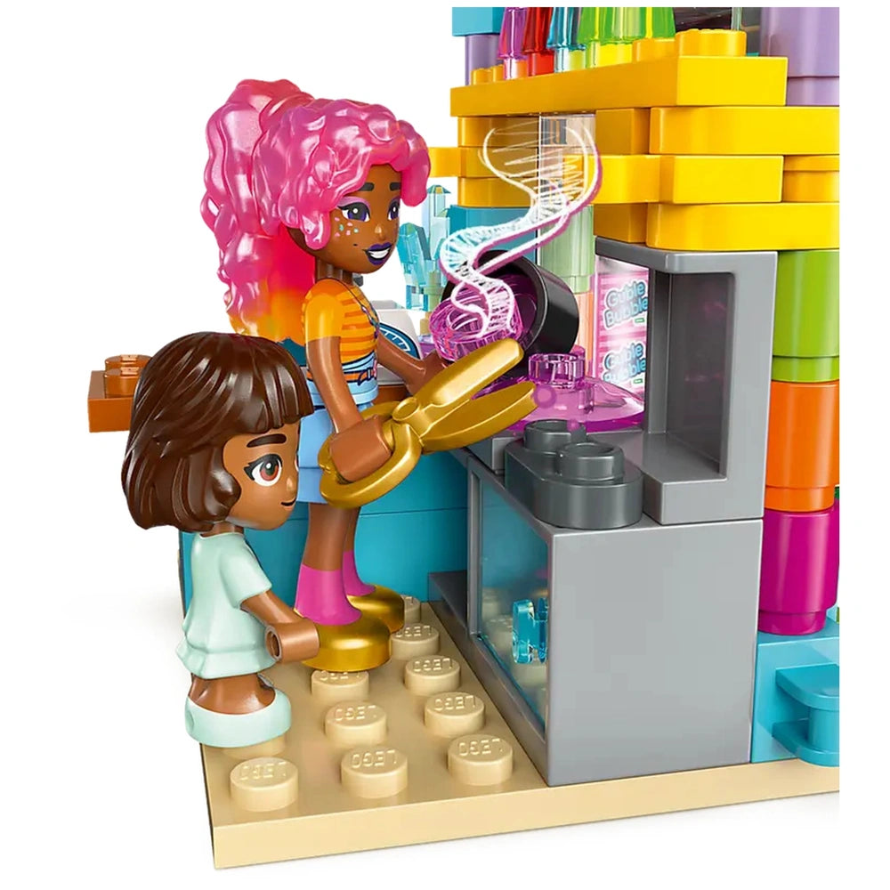 Two mini-dolls play at a vibrant LEGO candy store, surrounded by colorful candy jars, with one doll using scissors and the other watching eagerly.