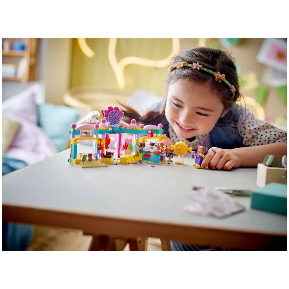 A smiling girl plays with the Lego Friends Heartlake City Candy Store set, featuring colorful storefront and mini-dolls on a light table.