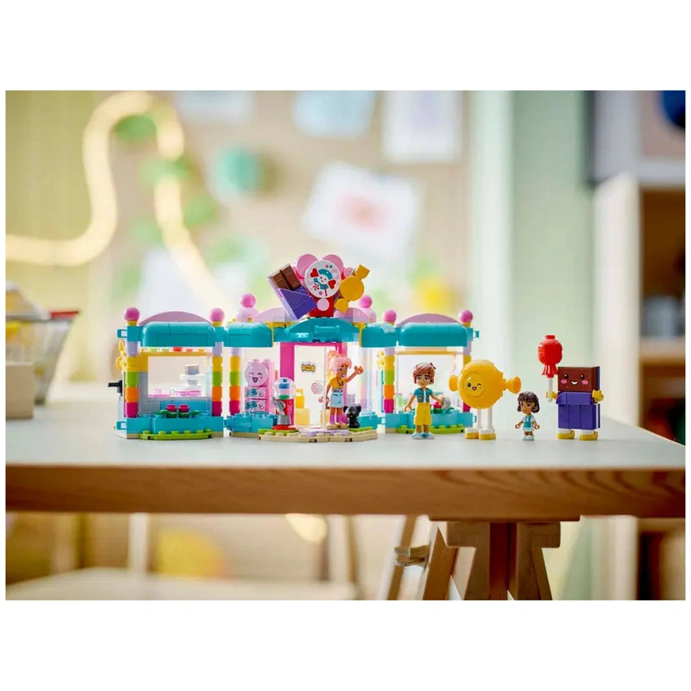 LEGO Friends Heartlake City Candy Store set featuring colorful buildings, mini-dolls, a playful cat figure, and candy-themed decorations on a wooden table.