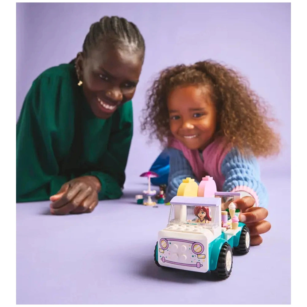 A smiling girl holds a colorful LEGO® Friends Heartlake City Ice Cream Truck with ice cream toppings. An adult smiles nearby, on a pastel background.