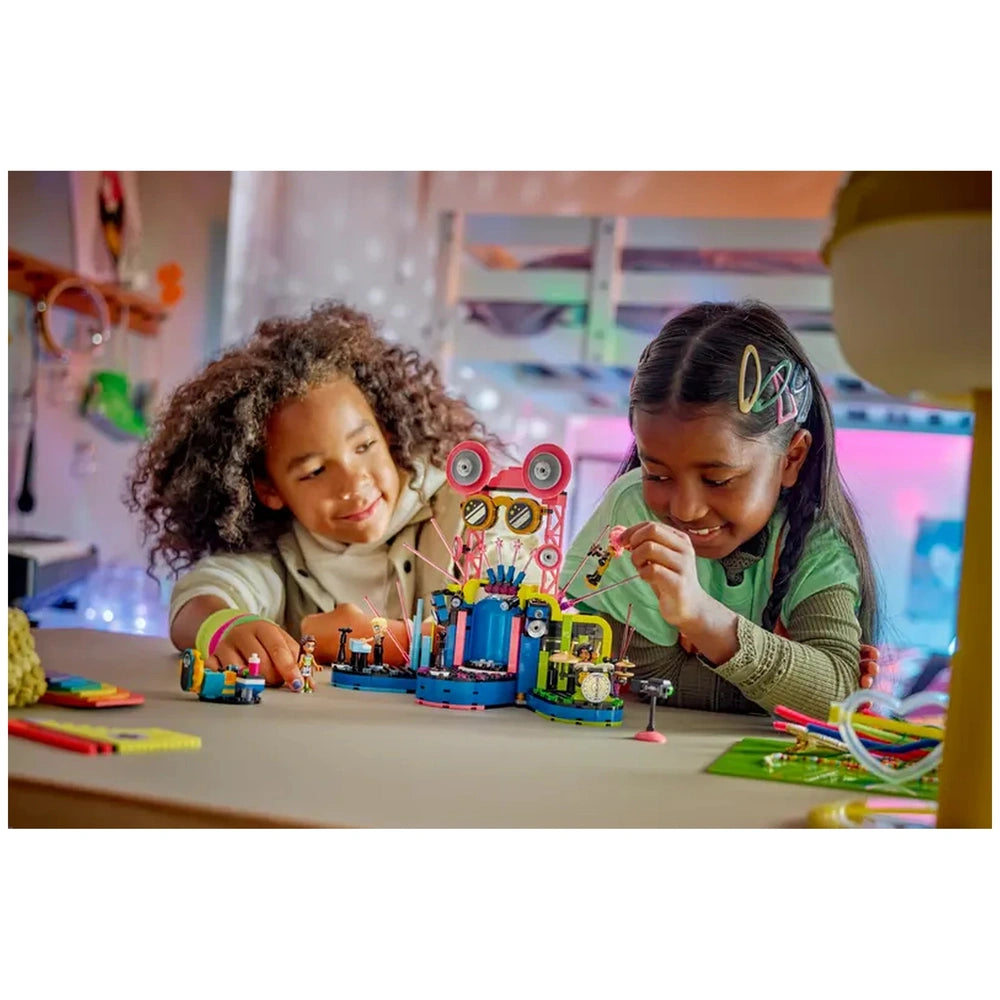 Two children play together with the LEGO® Friends Heartlake City Music Talent Show set, smiling as they arrange colorful pieces and accessories on a vibrant toy music stage, complete with speakers and a judge’s desk, in a cozy room filled with soft lighting.