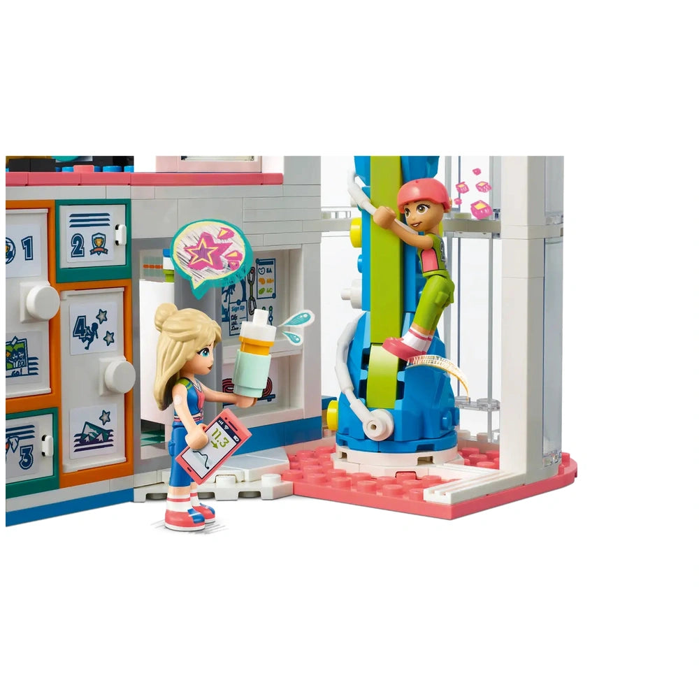 Two mini-dolls in a colorful LEGO® Friends Sports Center, one climbing a green wall and the other holding a drink and clipboard, engaged in playful activity.