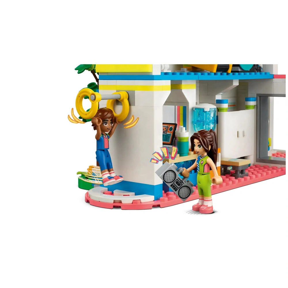 Two mini-dolls play at the LEGO® Friends Sports Center, one hanging from yellow rings while the other holds a boombox, showcasing a colorful and dynamic sports environment.