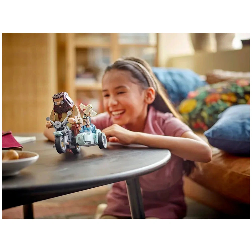 A young girl smiles joyfully as she plays with a LEGO construction set featuring Hagrid and Harry Potter's motorcycle, set on a table in a cozy living room.