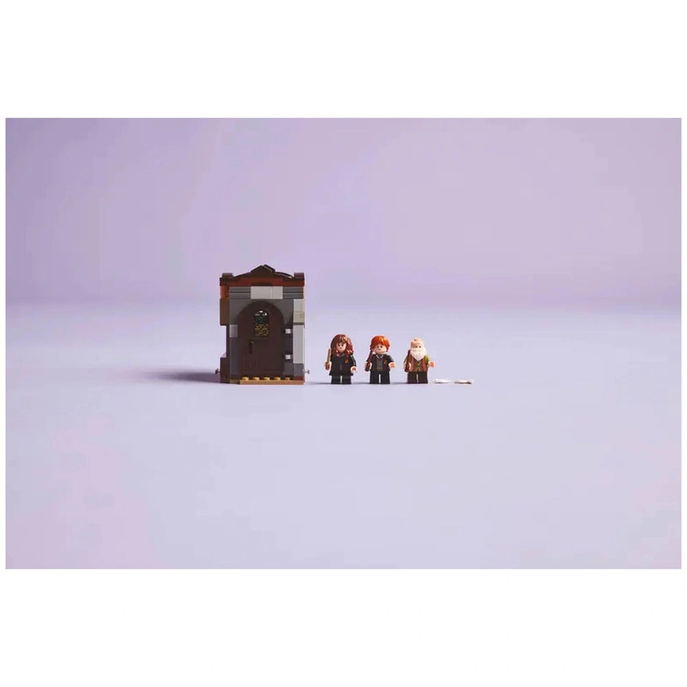 Three LEGO figures stand in front of a small Hogwarts classroom model. The figures include a young witch holding a wand, a red-haired wizard, and an elderly wizard. The classroom has a brown door and a colorful window, set against a soft, pastel purple background.