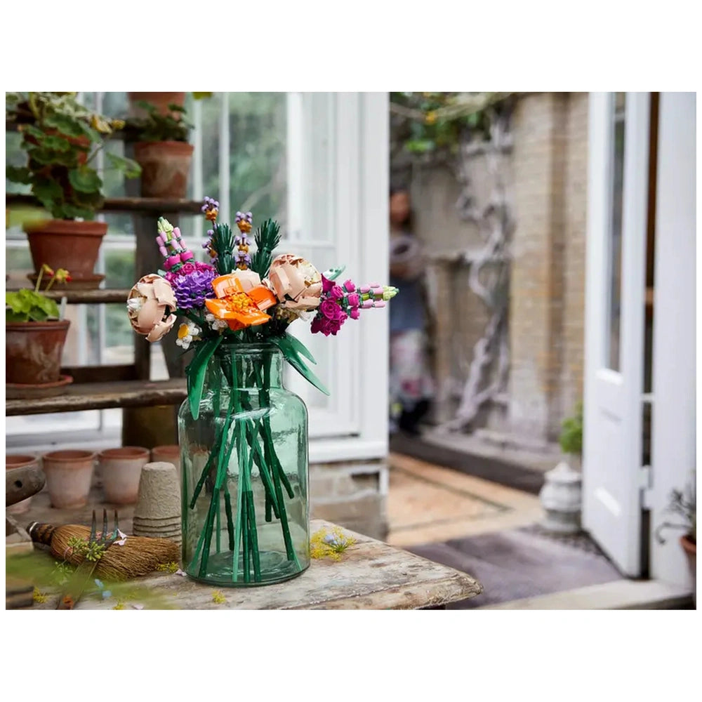 A vibrant LEGO Flower Bouquet featuring colorful flowers like orange roses, purple asters, and pink poppies in a clear glass vase, set on a rustic table near potted plants.