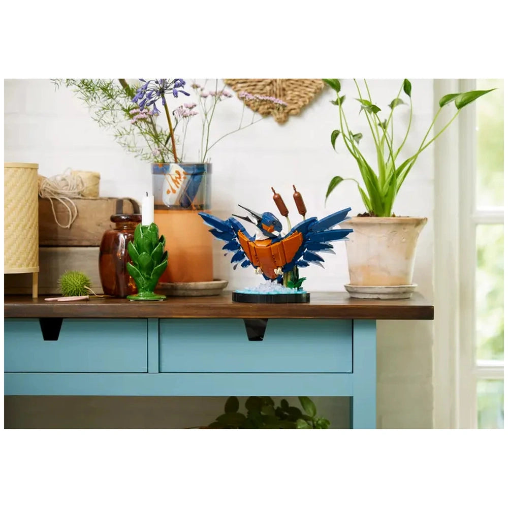 Colorful LEGO® Icons Kingfisher Bird construction set perched on a table, surrounded by green plants and decorative elements.