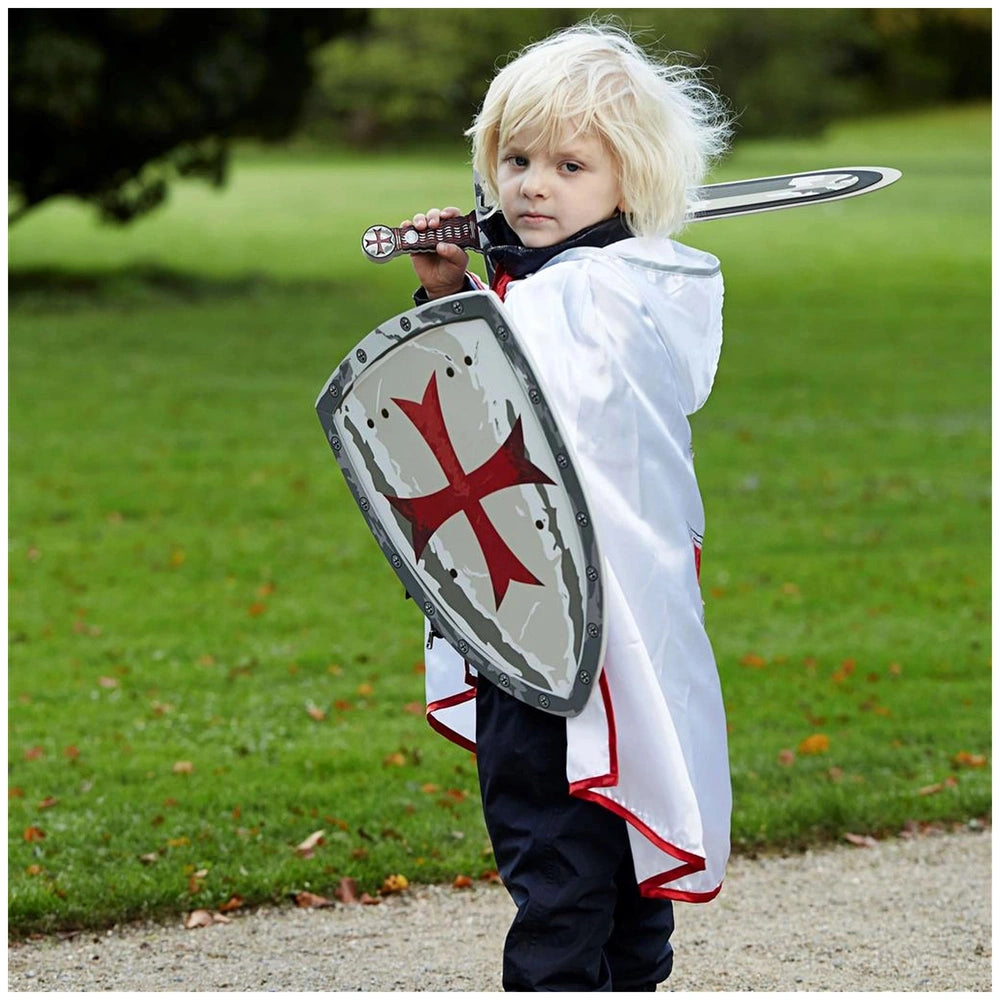 Blonde child in a Maltese Knight costume with a white jacket and red trim, holding a gray shield with a red cross on grass pa