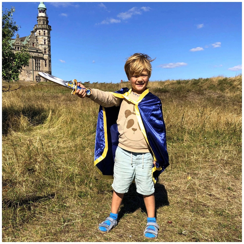 Mini Lion Sword in blue held by a child in a blue cape, brown shirt, and shorts, standing on dry grass near a stone building.