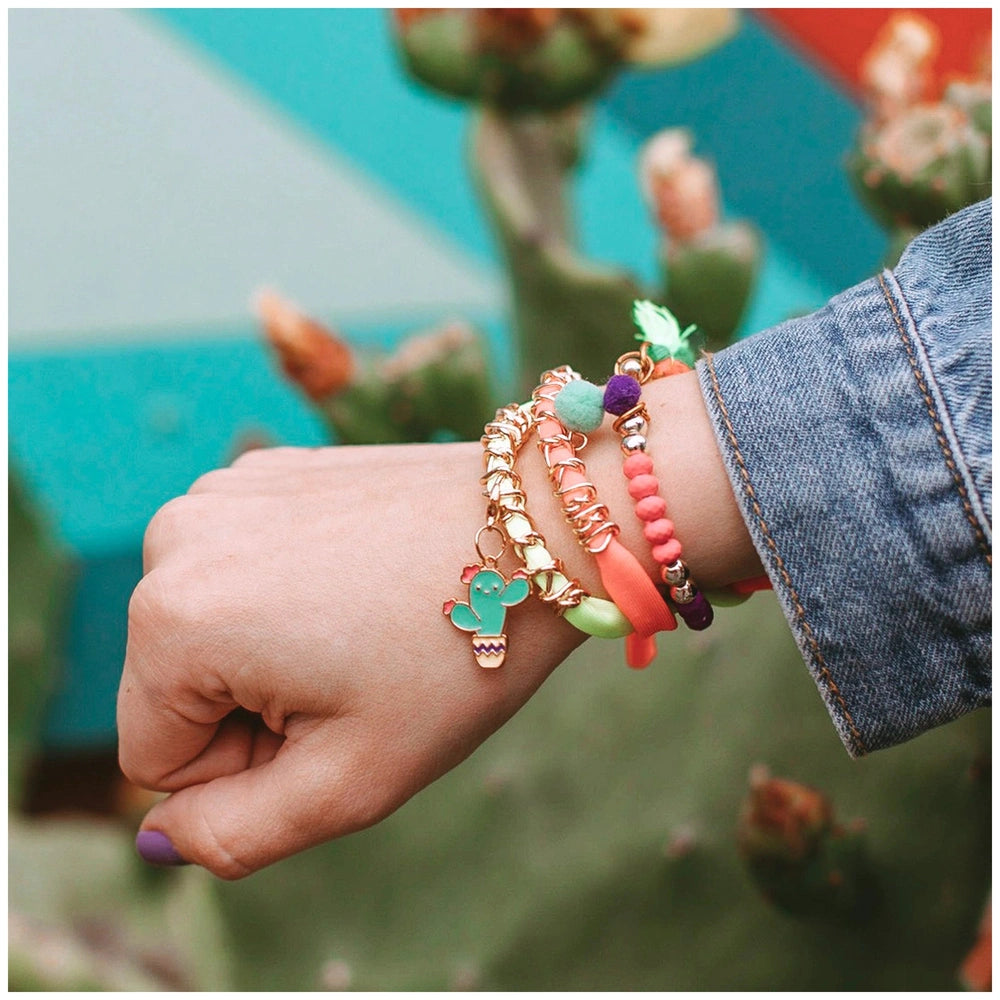 Neo-Brite chains and charms adorn a hand with painted nails, set against a blurred green background featuring a glossy cactus