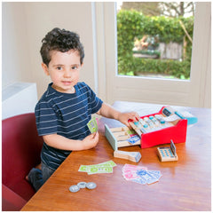 Young child in a blue striped shirt playing with a red cash register toy and green and white playing cards on a wooden table.