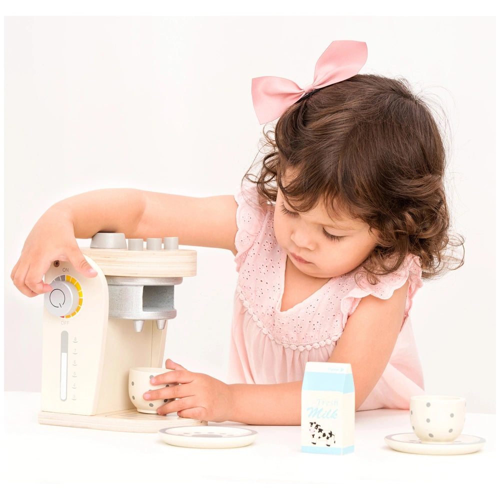 New Classic Toys coffee machine with a young girl in a pink polka dot dress, white cup, and milk carton on a white table.