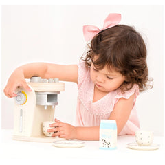 New Classic Toys coffee machine with a young girl in a pink polka dot dress, white cup, and milk carton on a white table.