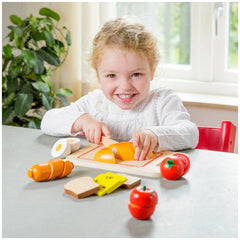 New Classic Toys breakfast cutting set with colorful plastic food items displayed on a wooden table, featuring a young girl p