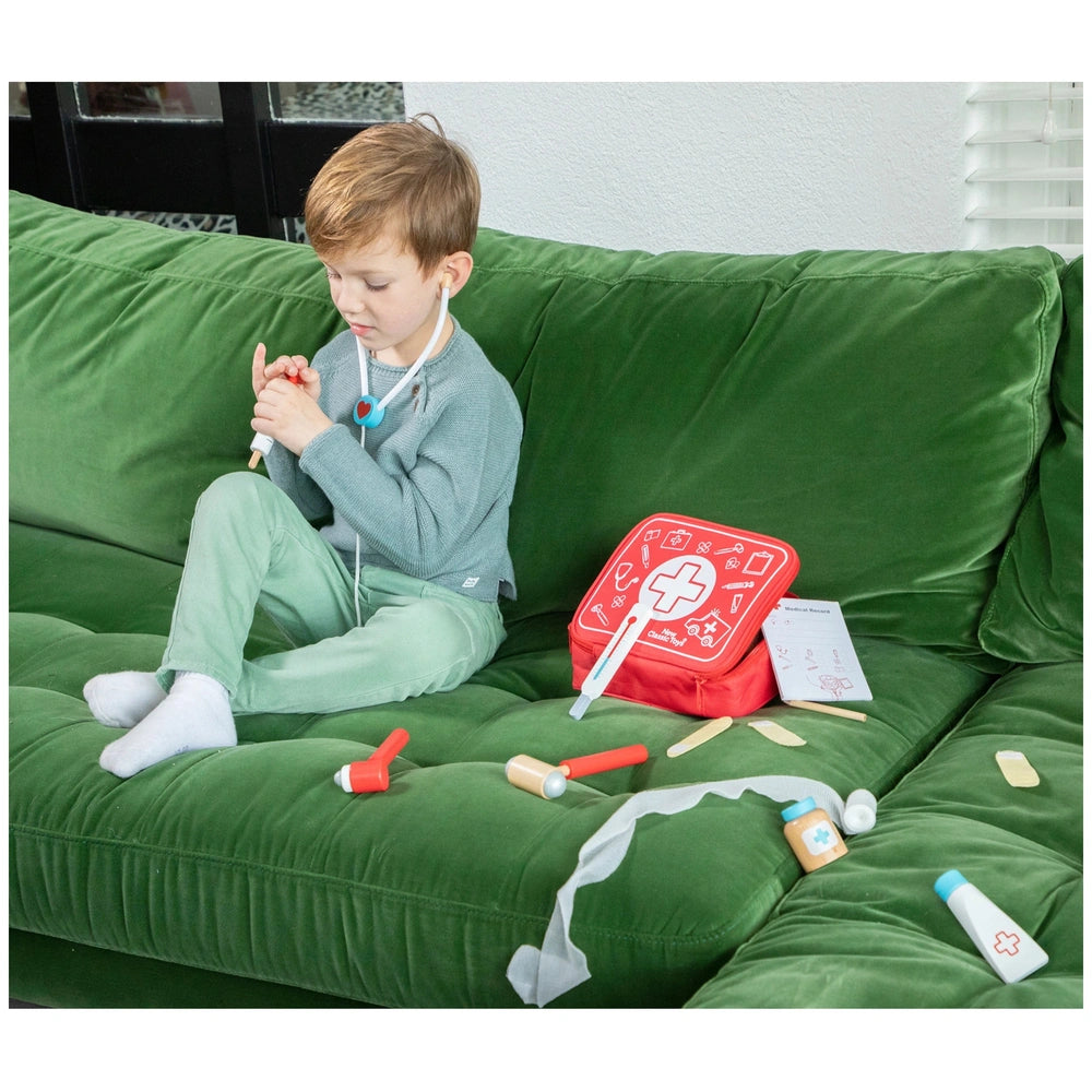 New Classic Toys doctor set features a child in a light green shirt and white headset, sitting on a green velvet couch.
