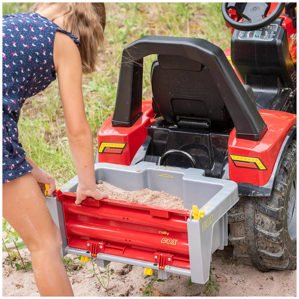Rollybox John Deere riding toy in red and black with a young girl reaching into its open cargo area on grass.