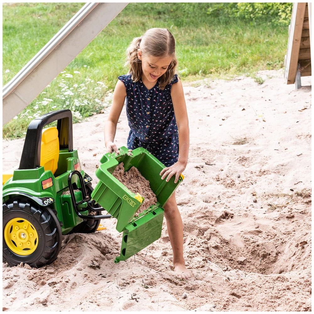 Rollybox John Deere green riding toy is held by a young girl in a blue floral dress playing in light brown sand.