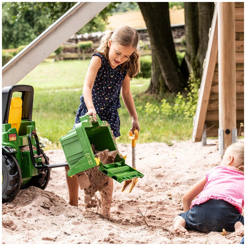 Rollybox John Deere Green riding toy sits in a sunny outdoor play area with a girl playing with a green shovel on sand.
