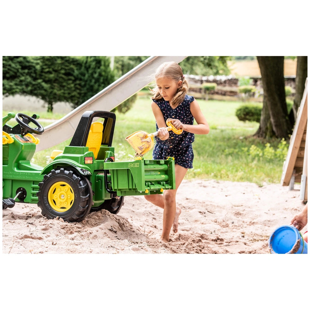 A young girl in a blue dress plays with a green and yellow toy tractor in a sandy outdoor play area.