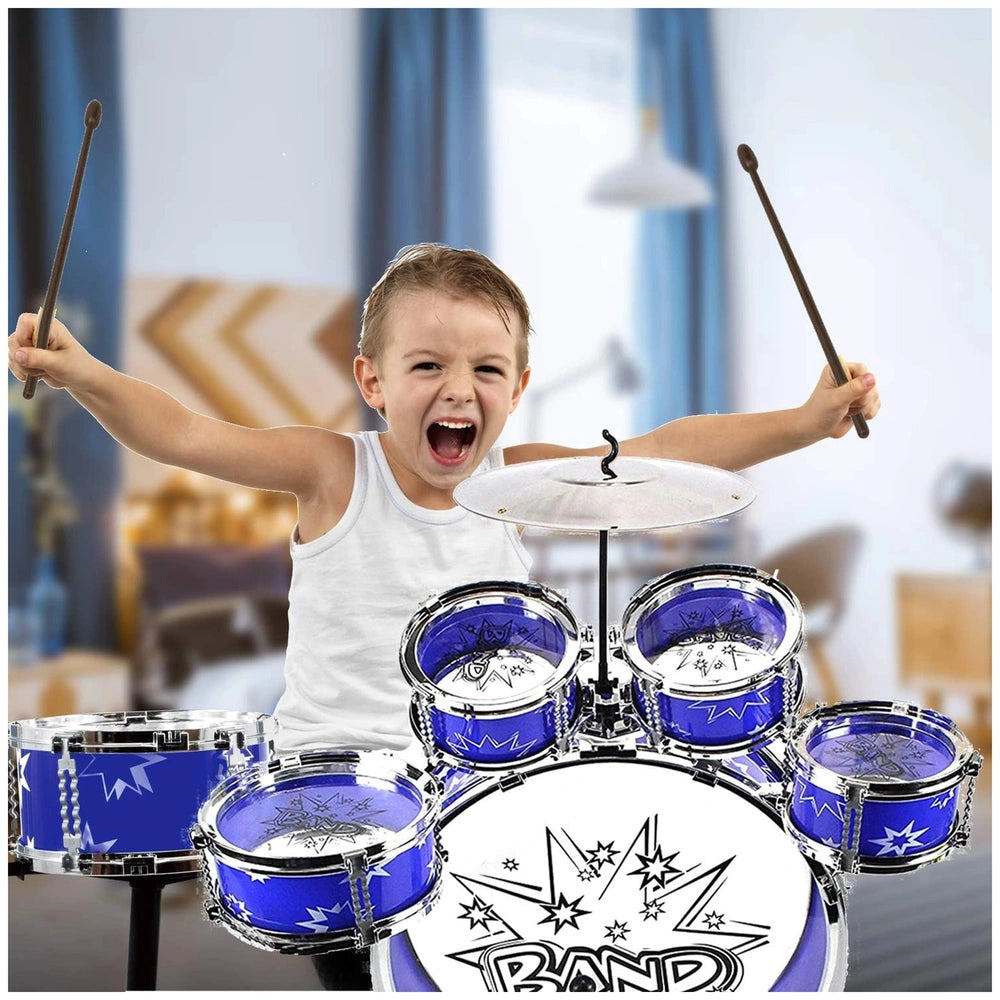 A young child in a white tank top plays a glossy metal drum set, energetically holding drumsticks with a blue curtain backdro