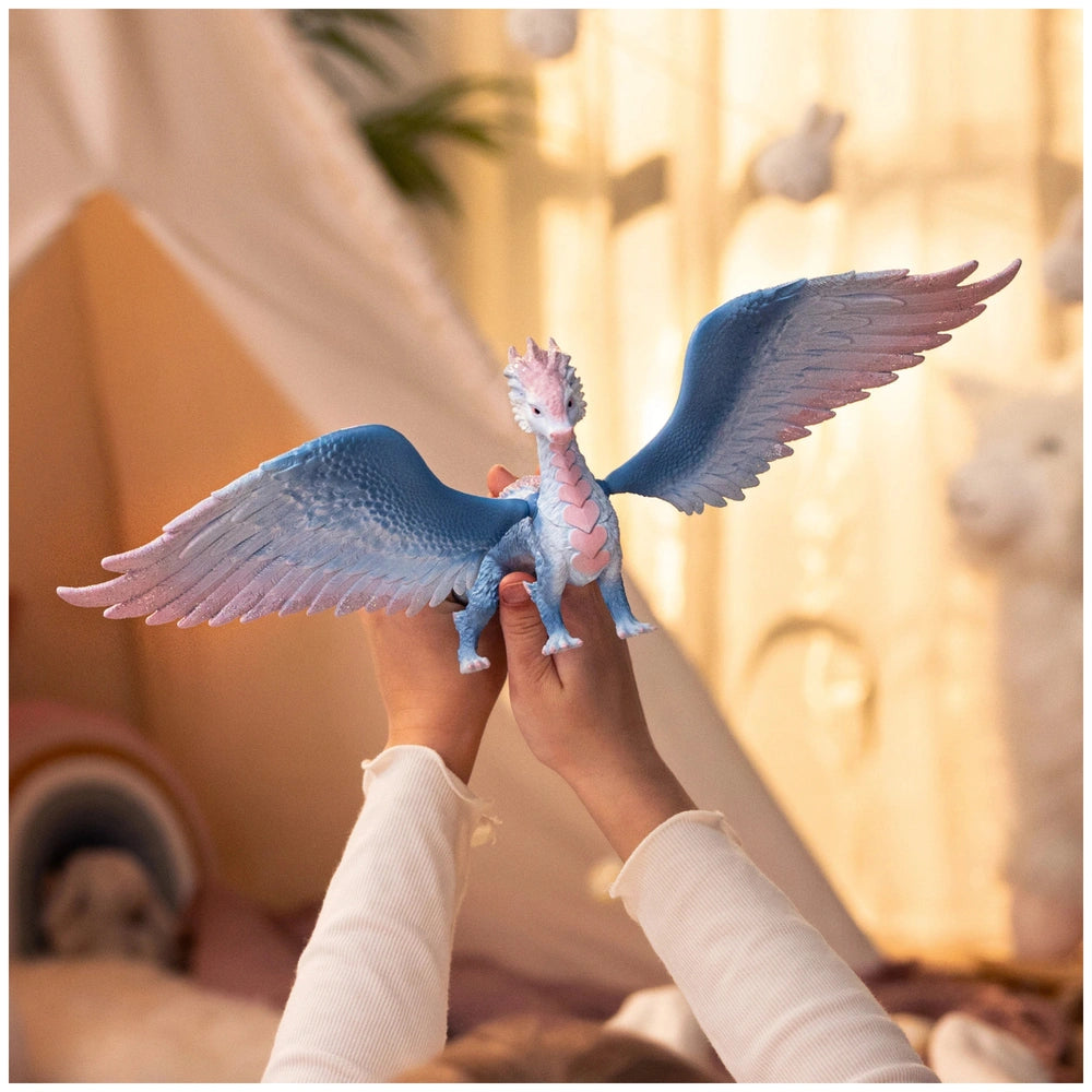 Blue crystal dragon toy with pink accents held between hands, set against a softly lit indoor backdrop.