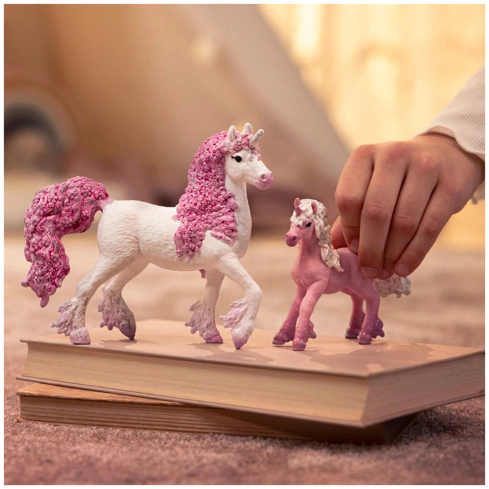Flower unicorn foal figurine in pink and white held by a hand, resting on a book in a softly lit indoor setting.