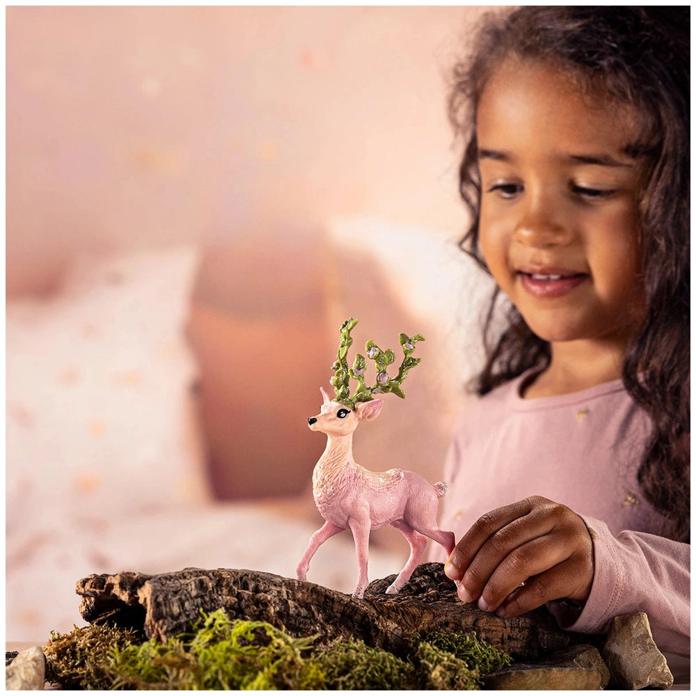 A young girl in a pink top smiles at a small pink toy deer resting on a moss-covered log in a cozy bedroom setting.