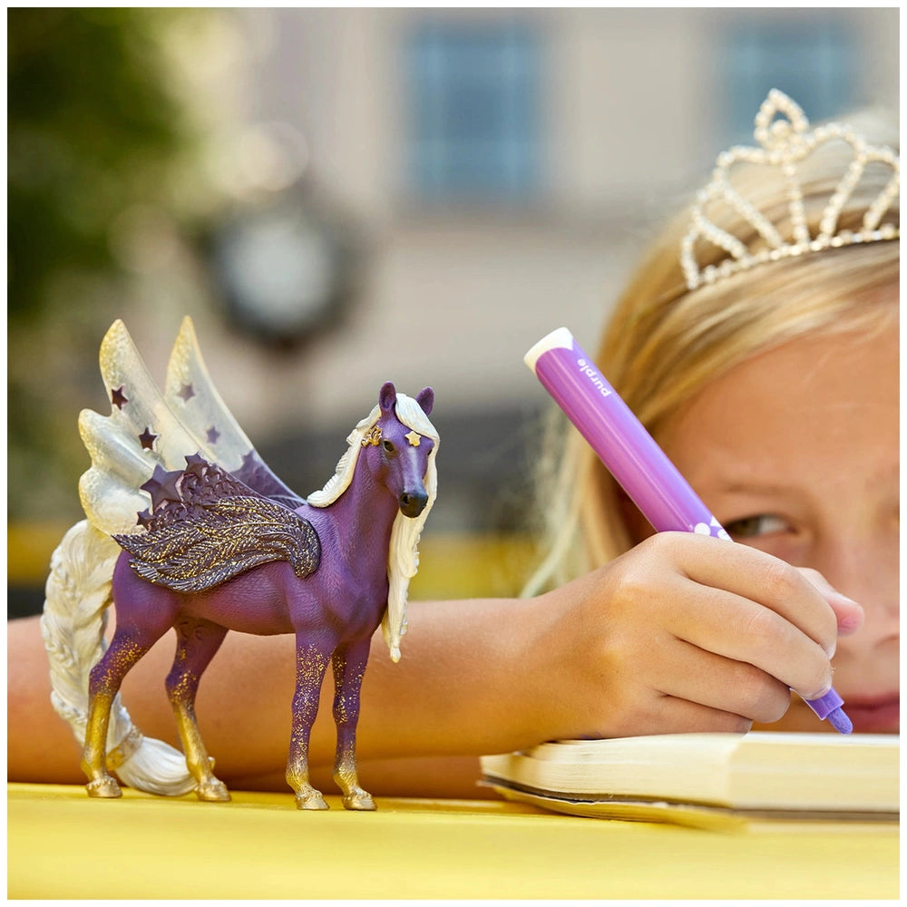 Schleich Star Pegasus Mare toy figure is featured alongside a young girl with a tiara, drawing with a purple pen at a yellow 