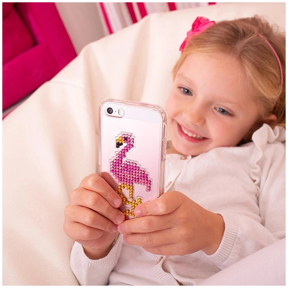 A young girl with blonde hair in a white top and pink headband smiles while holding a smartphone with a flamingo case.