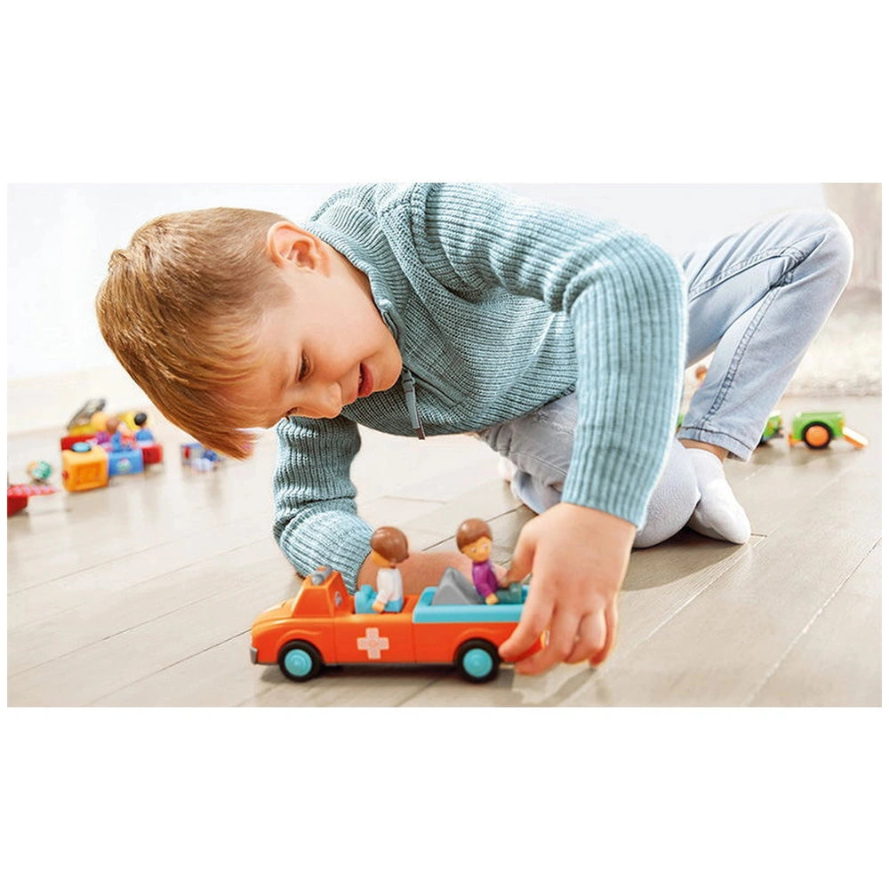 A young boy in a blue sweater and jeans plays with an orange toy truck on a wooden floor, leaning over it with excitement.