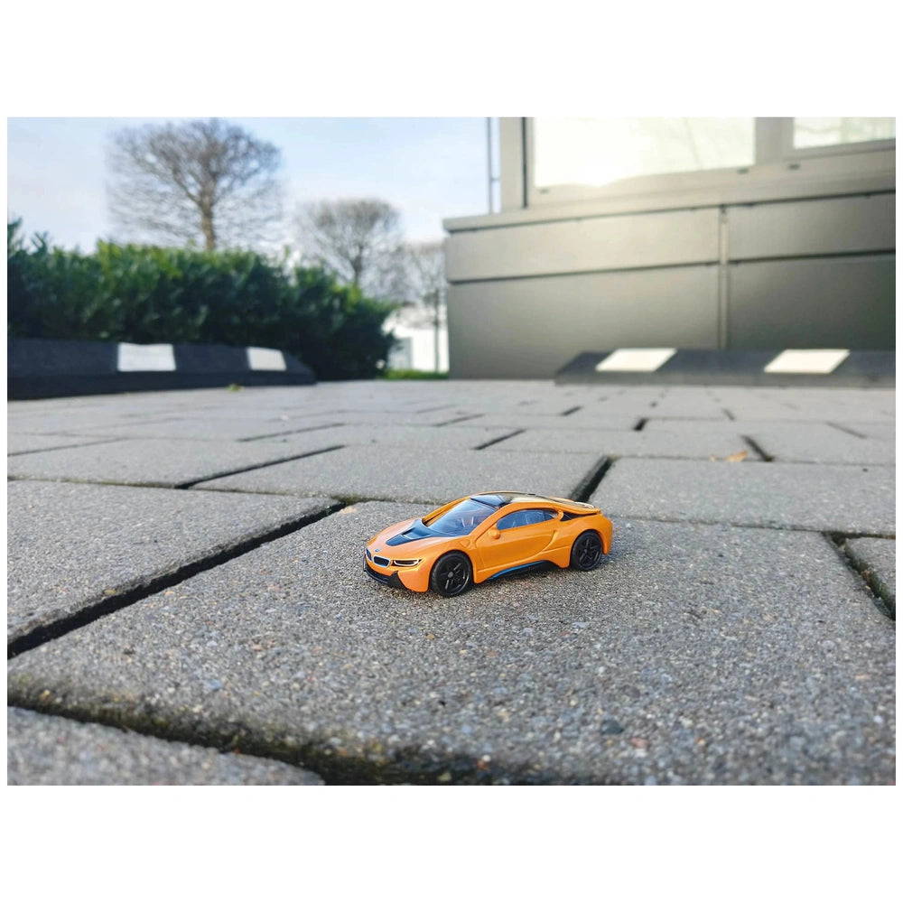 Siku BMW I8 model in orange with black accents, showcased on a sunny paved surface near a building and tree.