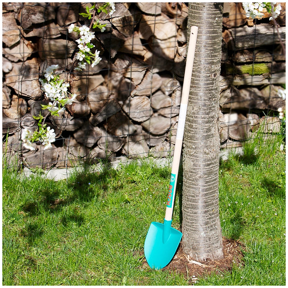 Theo Klein Bosch Garden Gardener Set with blue shovel on green grass near brown tree bark and white wooden fence.
