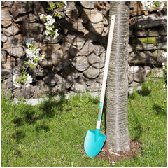 Blue plastic shovel placed on green grass in front of a brown wooden fence and a tree trunk with white flowers nearby.
